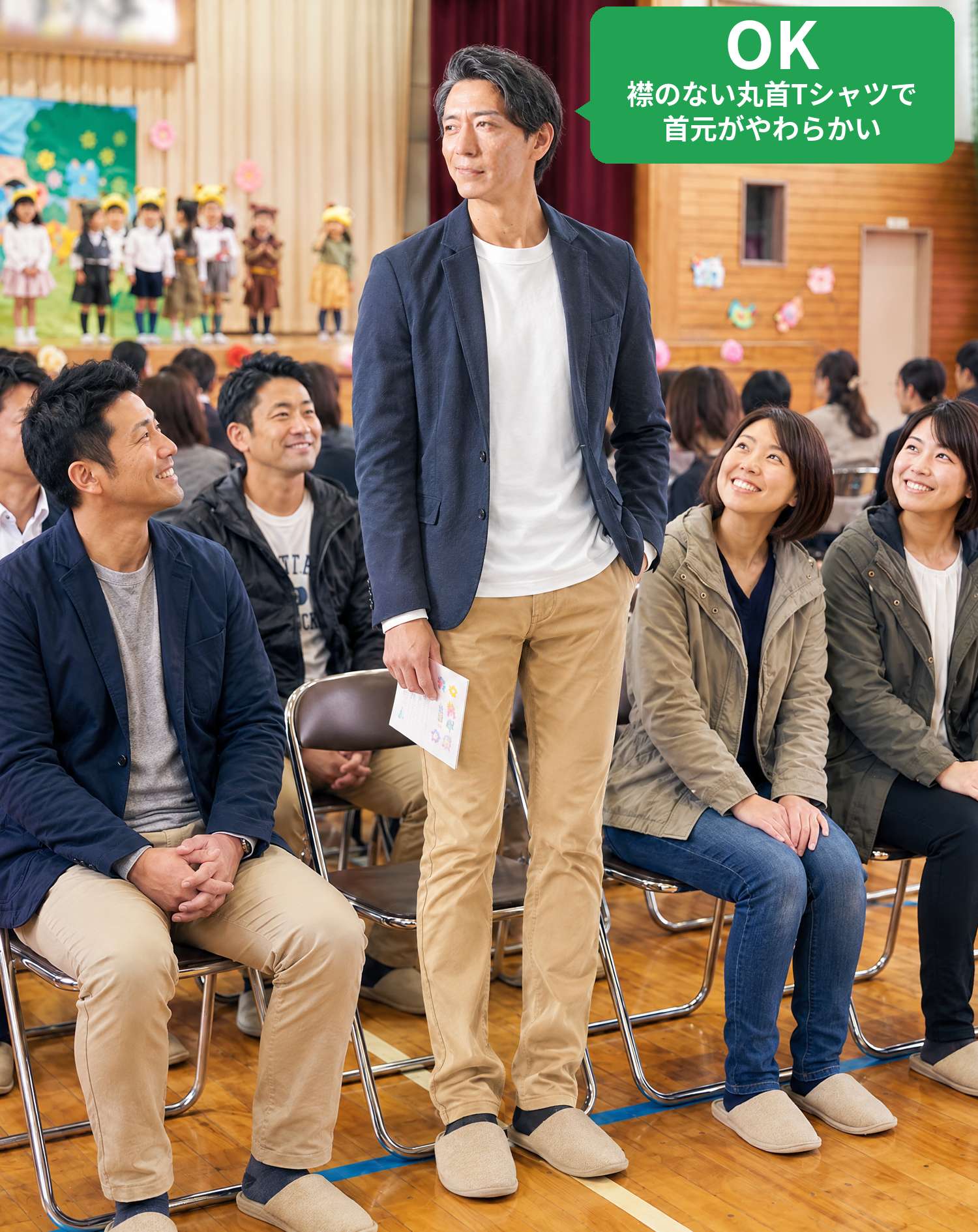 OKコーデ　体育館での子供の発表会で、客席のそばに立ち笑顔で舞台を見守るシーン（OKマーク付き）